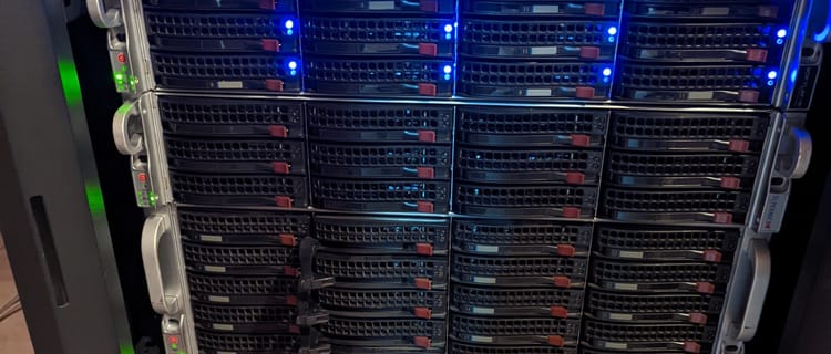 A Picture of multiple rack-mounted servers with hard-drive cages showing in a server rack with lights on indicating hard drives present, and power on